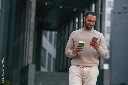 With cup of coffee and smartphone. Handsome black man is outdoors near the business building