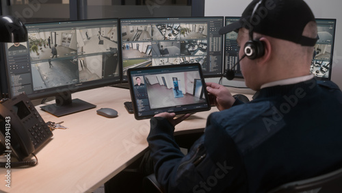 Security officer in headset controls and monitors CCTV cameras in coworking office, uses digital tablet and computers with playback on screens. High tech software showing surveillance cameras footage.