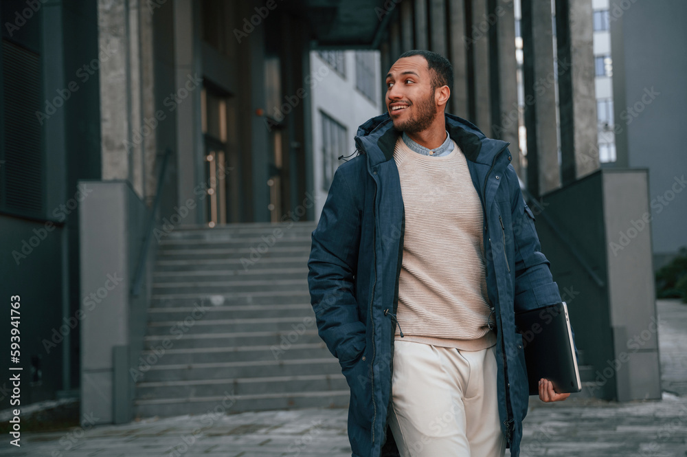 In blue jacket and with laptop in hands. Handsome black man is outdoors near the business building In blue jacket and with laptop in hands. Handsome black man is outdoors near the business building