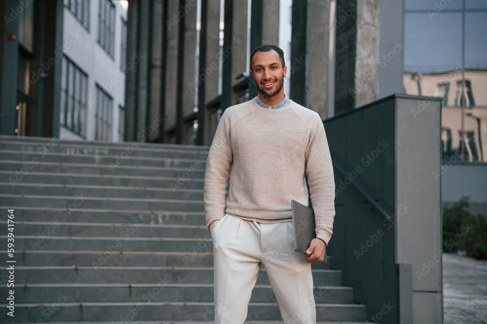 With laptop in hands. Handsome black man is outdoors near the business building