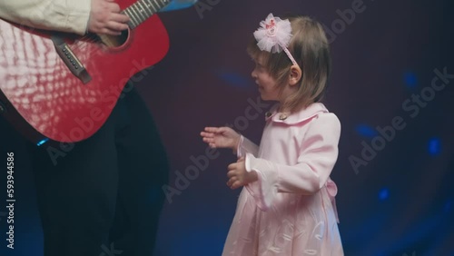 Little girl on stage in pink vintage dress, she dances, her father plays acoustic guitar. Color music is shining and smoke is billowing. Father Day. Performance on stage. Hobbies and creativity