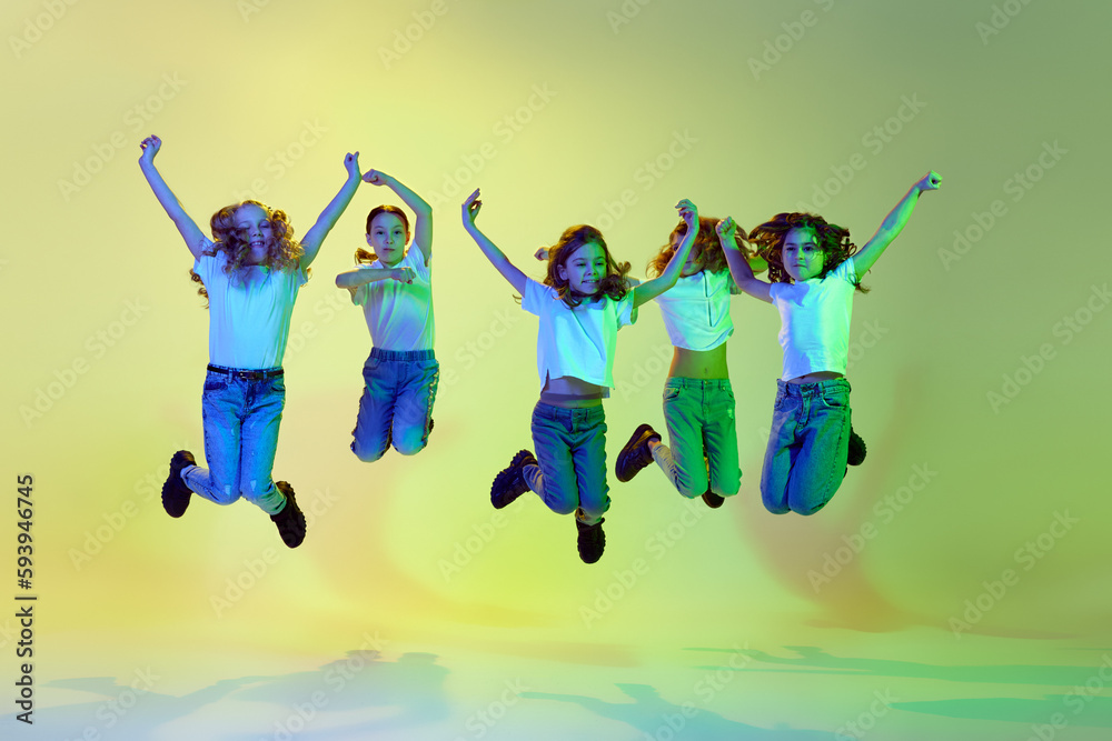 Cheerful, happy, little girls, children in casual sporty style clothes jumping, dancing against green studio background in neon light. Concept of childhood, hobby, sportive lifestyle, art, fashion