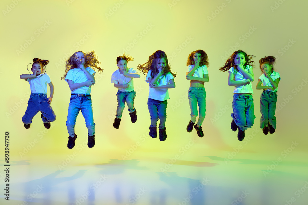 Performance. Little girls, children in casual sporty style clothes dancing, jumping against green studio background in neon light. Concept of childhood, hobby, sportive lifestyle, action and motion