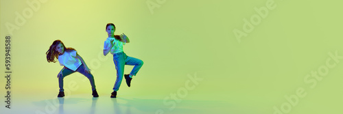 Active, talented little girls, children in casual clothes dancing against green studio background in neon light. Concept of childhood, hobby, sportive lifestyle. Banner. Cope space for ad