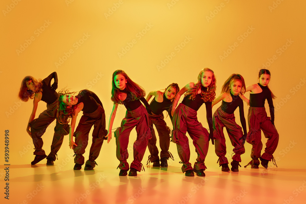 Expressive dance. Group of stylish little girls, kids, dancing, training hip-hop against yellow studio background in neon light. Concept of childhood, hobby, sportive lifestyle, education