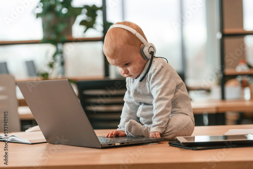 In headphones. Cute little baby boy is in the modern office on the table by laptop