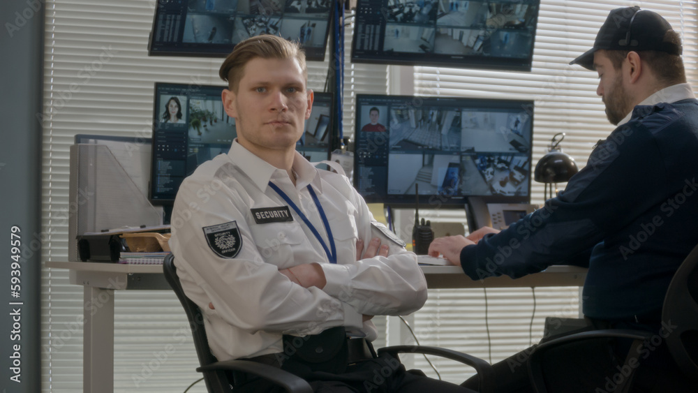 Security officer sits in observation room and looks at camera. Coworker monitors CCTV cameras footage on computer multiple screens at background. High tech software with AI facial recognition system. Security officer sits in observation room and looks at camera. Coworker monitors CCTV cameras footage on computer multiple screens at background. High tech software with AI facial recognition system.