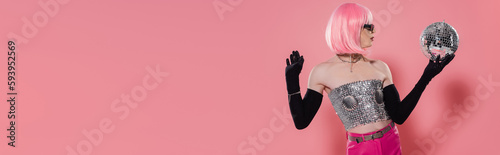 Fotografie Side view of stylish drag queen in shiny top and gloves holding disco ball on pink background, banner