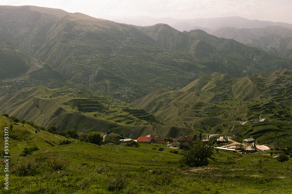 Majestic summer mountain landscape - bright green mountains ridges with lush green meadow terraces, village away in soft mist of early morning sunlight, panorama view. Amazing trip in Dagestan.