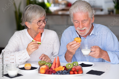 Cheerful senior couple at home having breakfast together with muffin, milk and fresh seasonal fruit, healthy eating concept