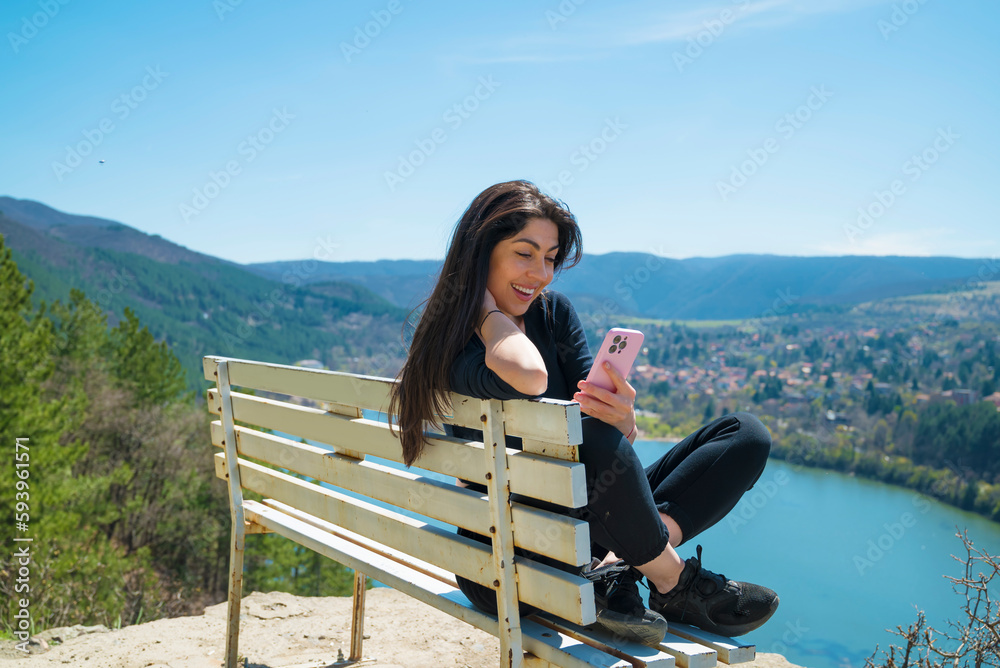 Beautiful woman sitting on a bench above a lake with stunning view looking her mobile phone Beautiful woman sitting on a bench above a lake with stunning view looking her mobile phone
