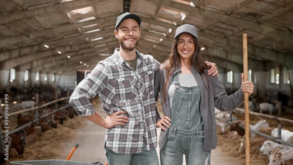 Zooming in pretty Caucasian couple with agricultural tools, standing in goats barn. Happy man hugging his wife and smiling. Young people taking care of farm animals together. Farming business concept. Zooming in pretty Caucasian couple with agricultural tools, standing in goats barn. Happy man hugging his wife and smiling. Young people taking care of farm animals together. Farming business concept.
