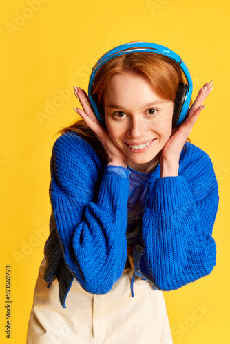 Portrait of young beautiful girl, teenager with red hair wearing headphones and smiling over yellow studio background. Happiness, joyful photo