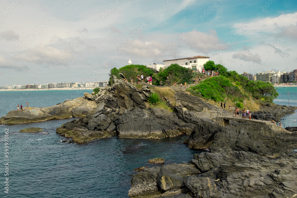 View of Praia do Forte, with the fort on top of the hill, sea entering amidst large rocks, cloudy sky and the city in the background.