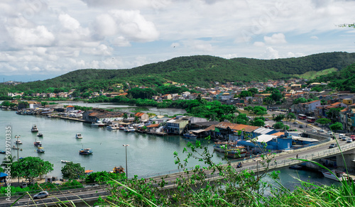 View of the port of Cabo Frio seen from the viewpoint of Nossa Senhora da Guia, with a bridge and many boats sailing through the channel, mountains around and a beautiful blue sky with clouds.