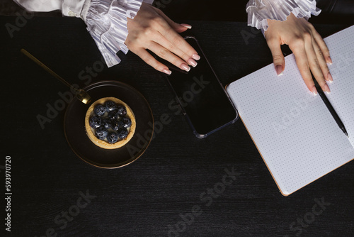 Black wooden desk with a notebook, phone, small blueberry cake and a woman's hands in a fancy shirt. flat lay. top view.