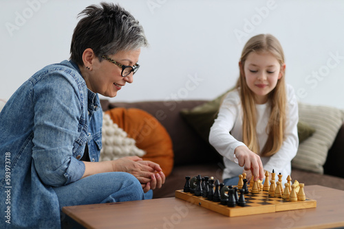Mature woman in glasses plays chess with a child girl in a home interior.