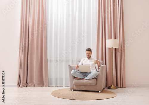 Smiling man drinking tea while working on laptop near window with beautiful curtains at home. Space for text
