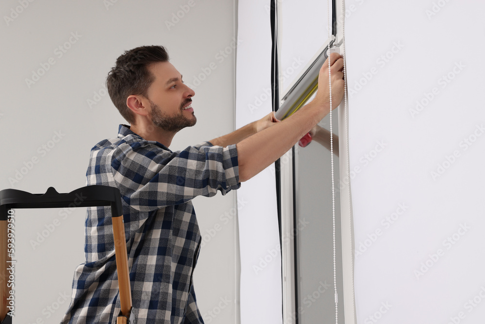 Man measuring window with tape on stepladder indoors. Roller blinds installation Man measuring window with tape on stepladder indoors. Roller blinds installation