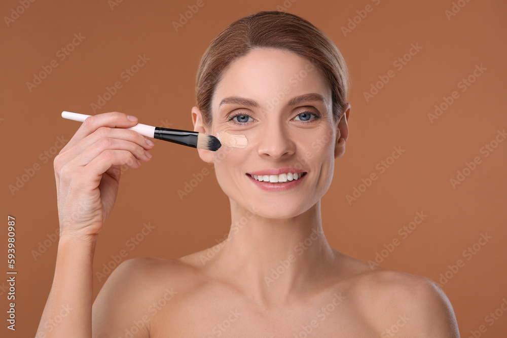 Woman applying foundation on face with brush against brown background Woman applying foundation on face with brush against brown background