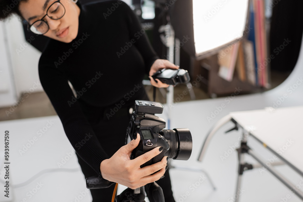 blurred african american content manager adjusting digital camera while holding exposure meter in photo studio.