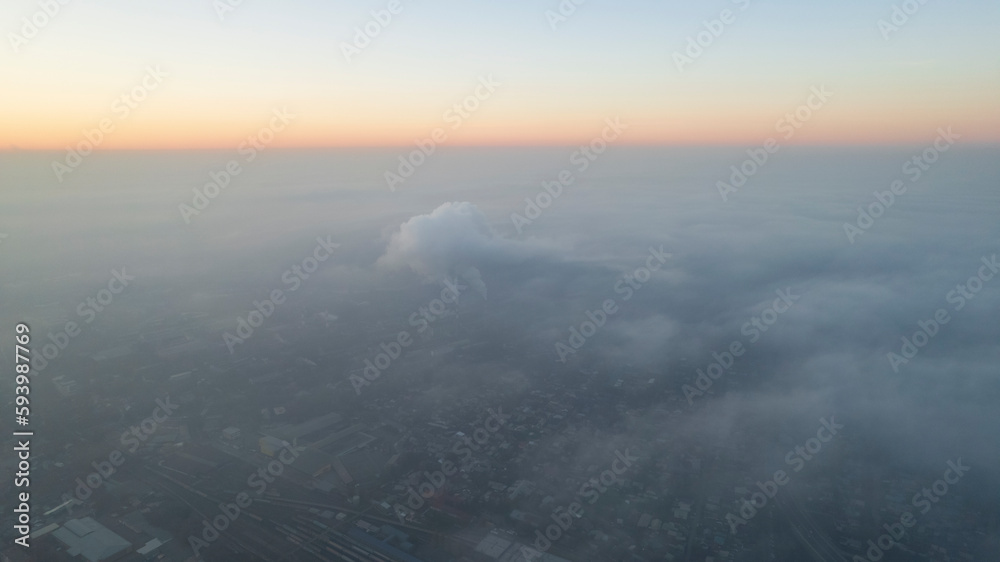 Epic gray smog is visible at sunset over the city. Aerial view from the drone of houses, roads, cars and parks. White clouds are illuminated by orange rays of the sun. The city is suffocating. Almaty