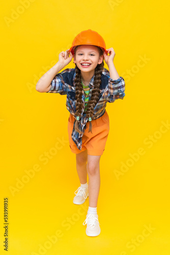A little girl in an orange construction helmet is holding her head and smiling broadly, happily. The child is preparing for repairs in the nursery. In a mouthful. Vertical. Yellow isolated background