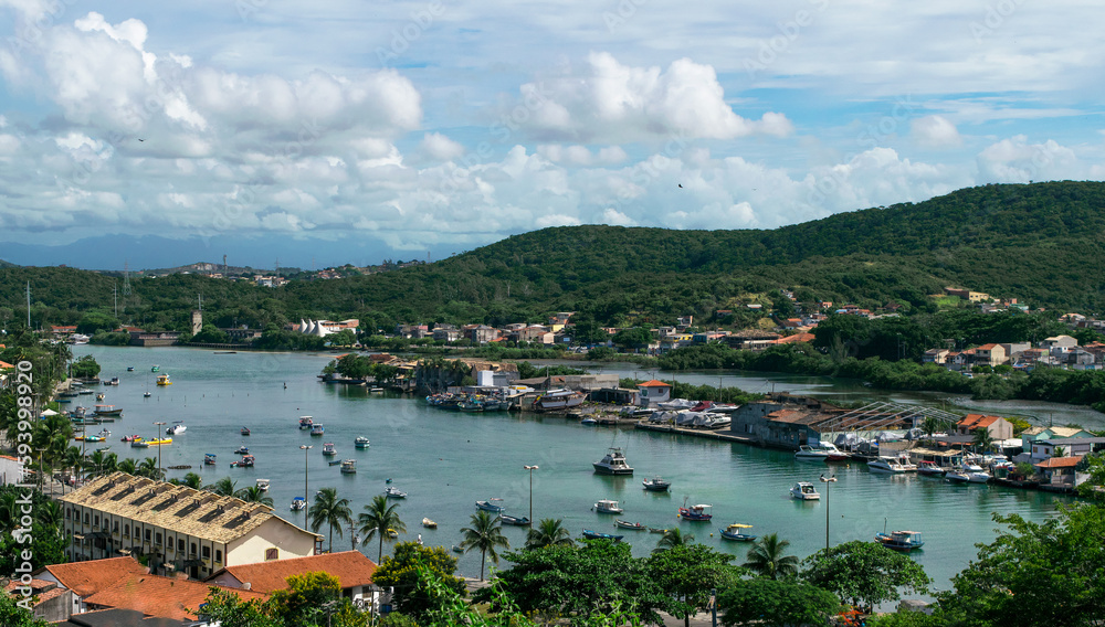 Small port of Cabo Frio seen from the viewpoint of Nossa Senhora da Guia, many boats anchored and others sailing through the channel, mountains around and beautiful blue sky with clouds.