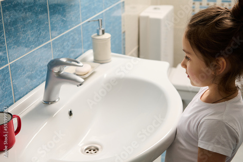 Side portrait of a beautiful 5 years old Caucasian little child girl in white pajamas, standing by sink in the bathroom during daily morning hygiene routine. Childhood. Lifestyle. Domestic life