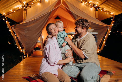 Happy family with lovely baby relaxing and spend time together in glamping on summer evening near cozy bonfire. Luxury camping tent for outdoor recreation and recreation. Lifestyle concept