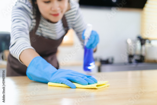 Asian housewife sanitizing surfaces cleaning home kitchen table with disinfectant spray bottle washing surface with towel and gloves.