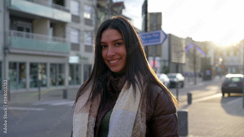 Fototapeta premium Portrait of a beautiful young woman standing in city street during sunset time with sunlight flare shining. Happy expression of a brunette adult girl in 20s smiling at camera