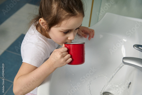 Close-up portrait of 5-6 years old Caucasian little child girl in white pajamas, rinsing her mouth with water while brushing teeth, standing by ceramic sink in the home bathroom. Oral care and hygiene