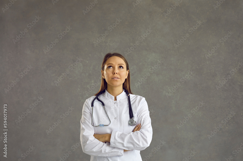 Young female doctor or nurse thinking of an interesting idea. Portrait of a pensive woman in a white lab coat looking up standing isolated on a grey background. Professional healthcare concept Young female doctor or nurse thinking of an interesting idea. Portrait of a pensive woman in a white lab coat looking up standing isolated on a grey background. Professional healthcare concept