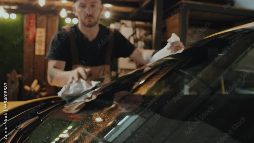 Tight medium shot of tattooed auto mechanic in overalls and black cap backwards washing car windscreen with spray and cloth in garage. Several light bulbs in background
