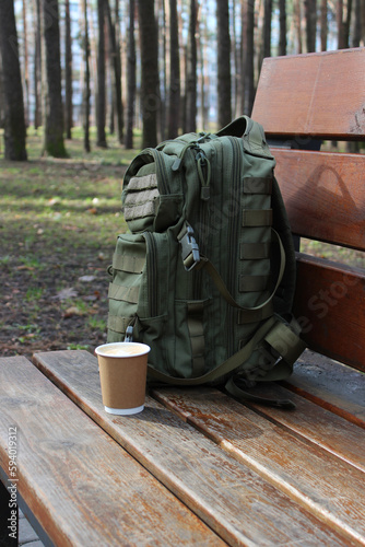 A cup of coffee next to a backpack leaning against the back of a wooden bench 