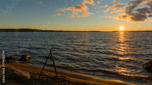 A photography tripod on a beach with a sunset behind a lake in summer in Finland