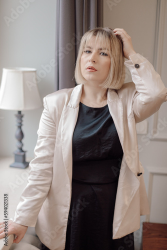 Beautiful blue-eyed blonde with square hairstyle looks at camera, standing near window in her office. Success in work. 