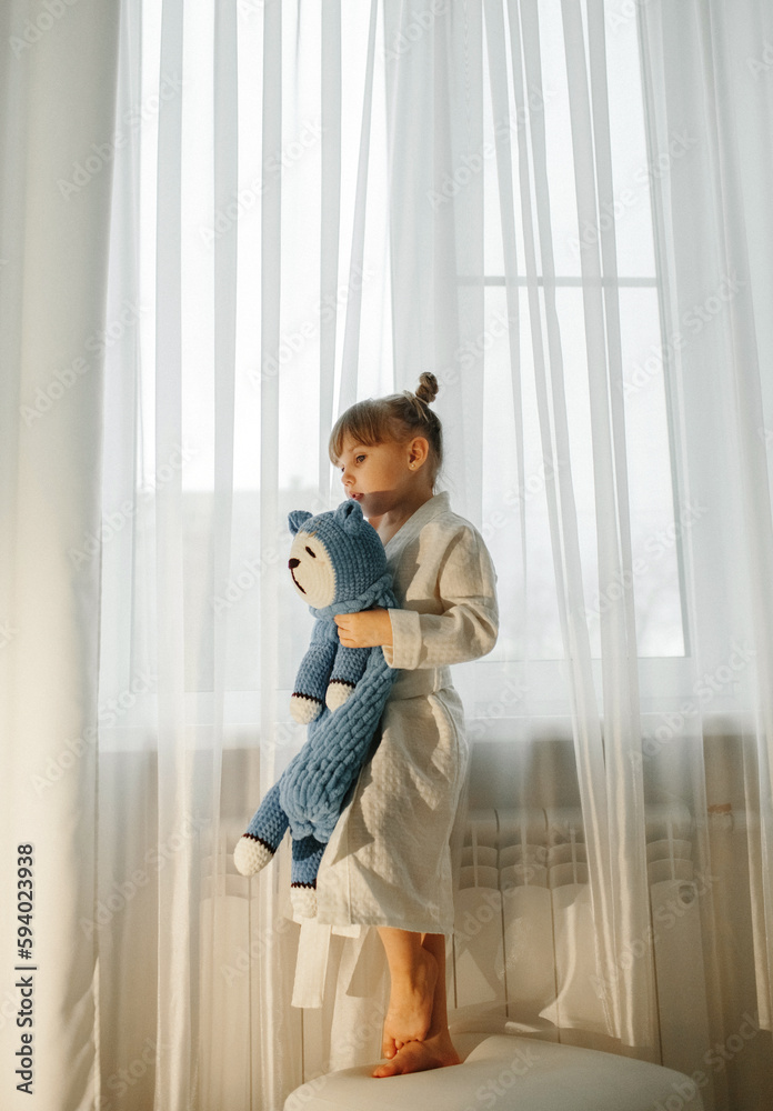 A girl in a dressing gown stands by the window and plays with a knitted toy A girl in a dressing gown stands by the window and plays with a knitted toy