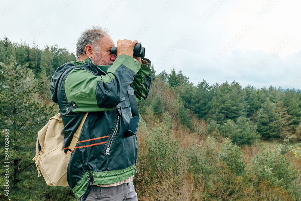 Fototapeta premium Mature bearded traveler man hiking in mountain forest looking with binoculars panoramic mountain valley.