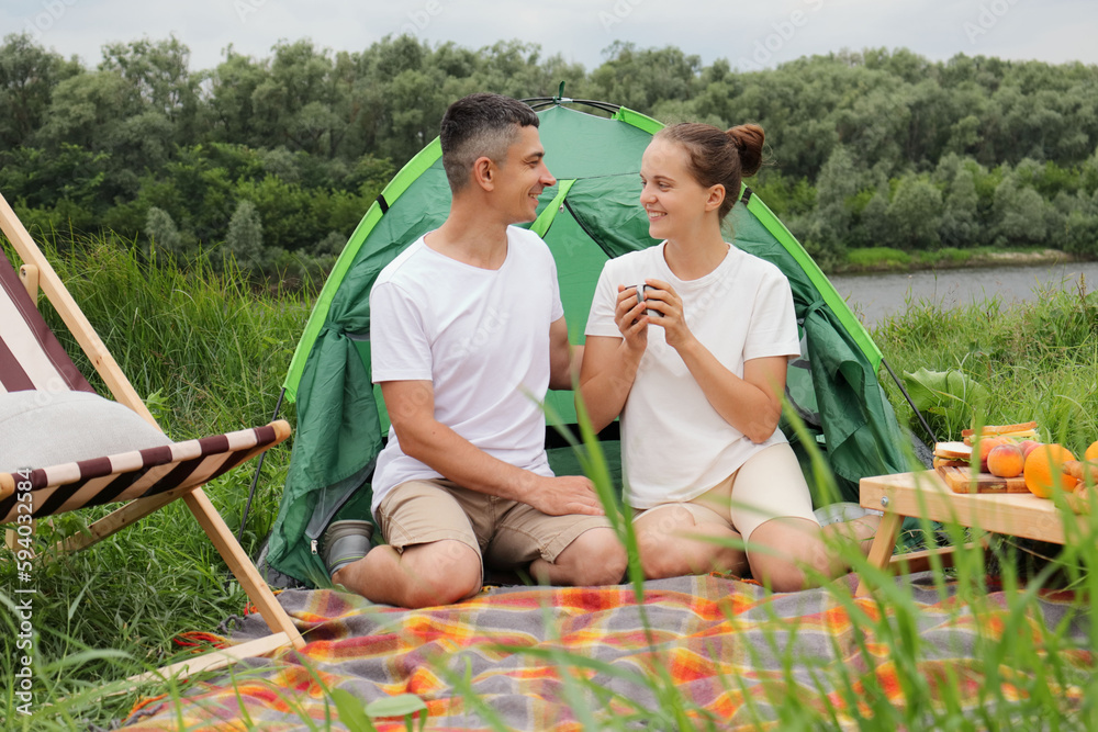 Vacation in nature. Loving woman and man wearing casual clothing sitting at tent near the river, enjoyoing hot coffee or tea, looking at each other, smiling happily. Vacation in nature. Loving woman and man wearing casual clothing sitting at tent near the river, enjoyoing hot coffee or tea, looking at each other, smiling happily.