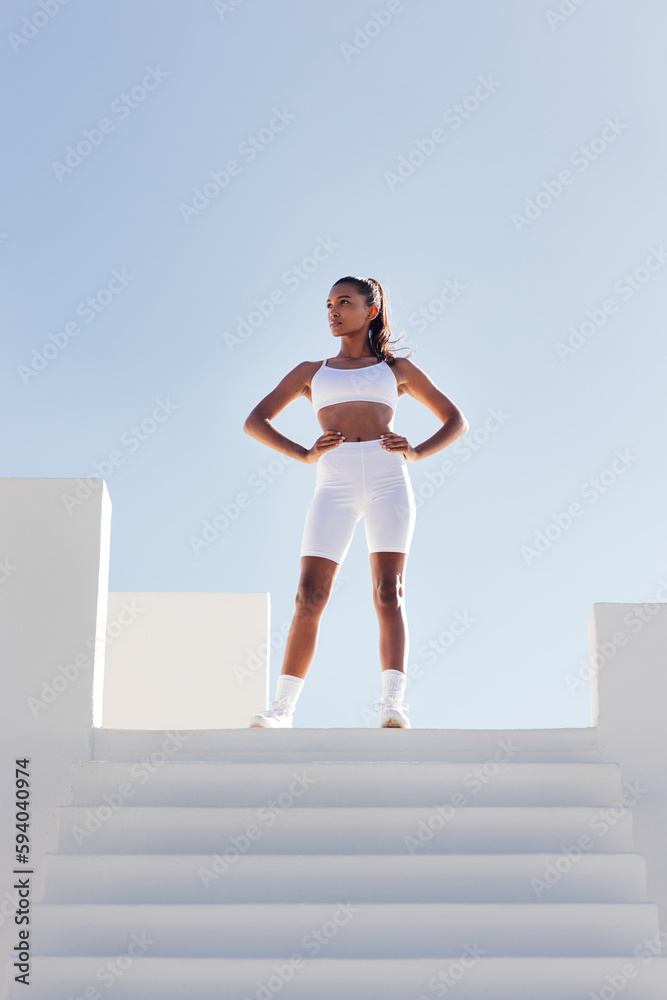 Full length of a slim confident woman looking away while standing on the top of stairs. Sportswoman in white fitness attire against skies.