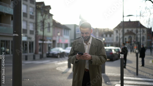 One happy Moroccan young man walking in city street looking at cellphone smiling. A male Middle Eastern person walks in sidewalk during sunset afternoon with flare