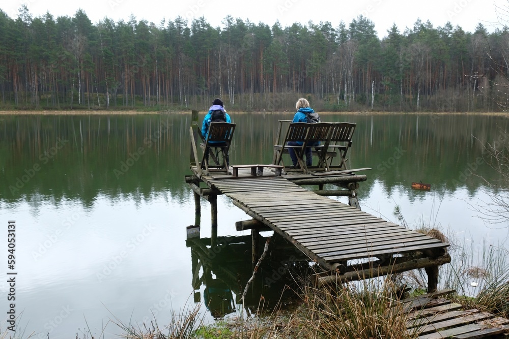 Obraz premium Two women in blue jackets are sitting on a wooden pier by the river on a spring day.