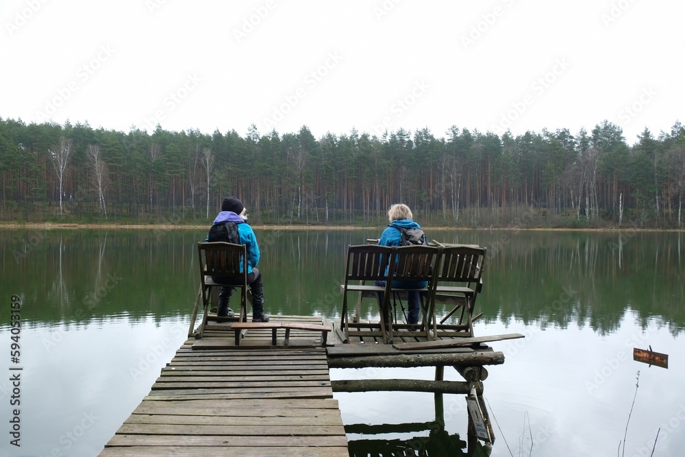 Fototapeta premium Two women in blue jackets are sitting on a wooden pier by the river on a spring day.