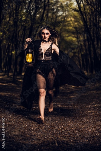 Vertical shot of a gothic girl in a transparent dress and black cape holding a lantern in forest
