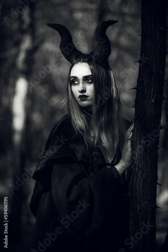 Grayscale of a gothic girl with Maleficent horns and black cape leaning to a tree with thorns