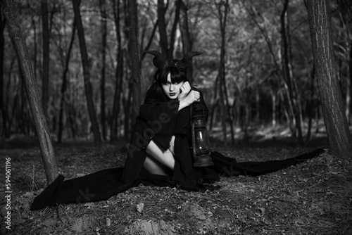 Grayscale of a young girl with Maleficent horns and black cape in a forest holding a lantern