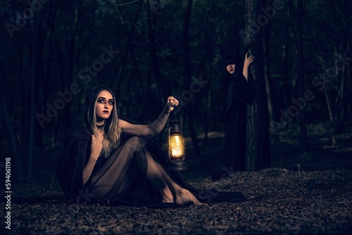 Night view of young gothic girls in black dresses holding a vintage lantern in a forest