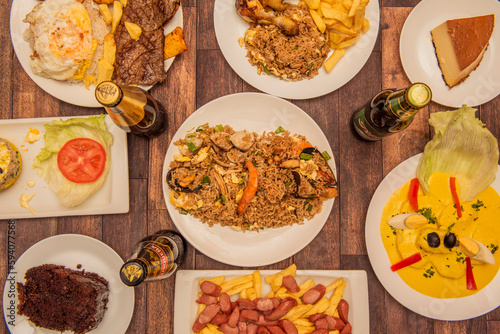 A set of Peruvian food dishes with papas a la huancaina in the center, a quesillo, steaks with rice and plantains, arros chaufa and drinks on a brown table photo